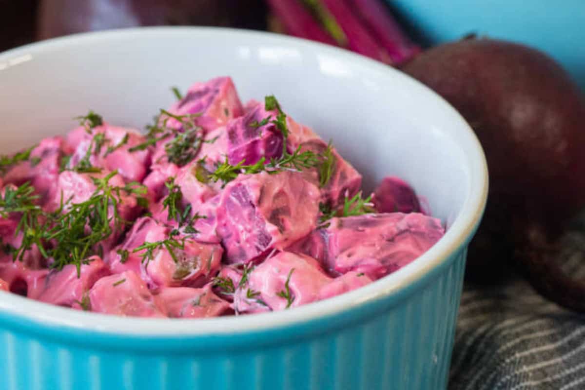A blue bowl filled with a creamy beet salad garnished with chopped dill with whole beets visible in the background.
