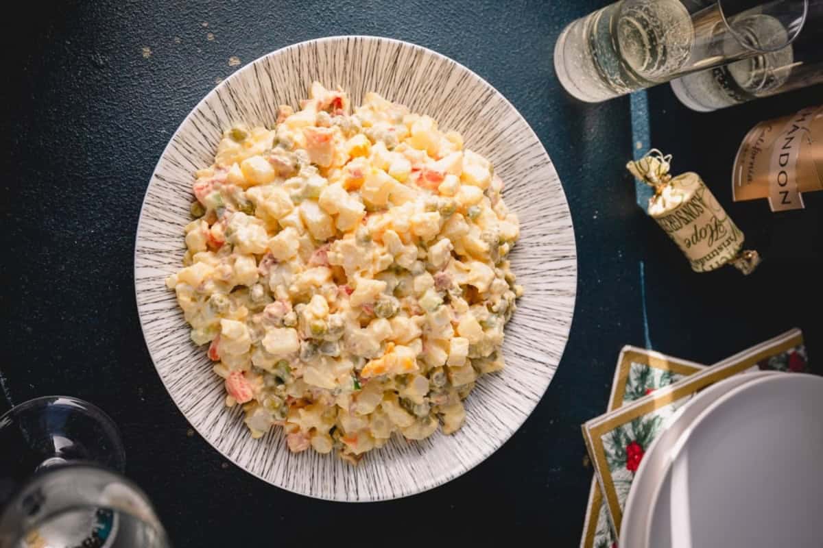 A plate of Russian Olivier salad with diced potatoes, vegetables, meat, hard boiled eggs and mayonnaise, sits on a table next to two glasses of sparkling beverage and a small wrapped gift.