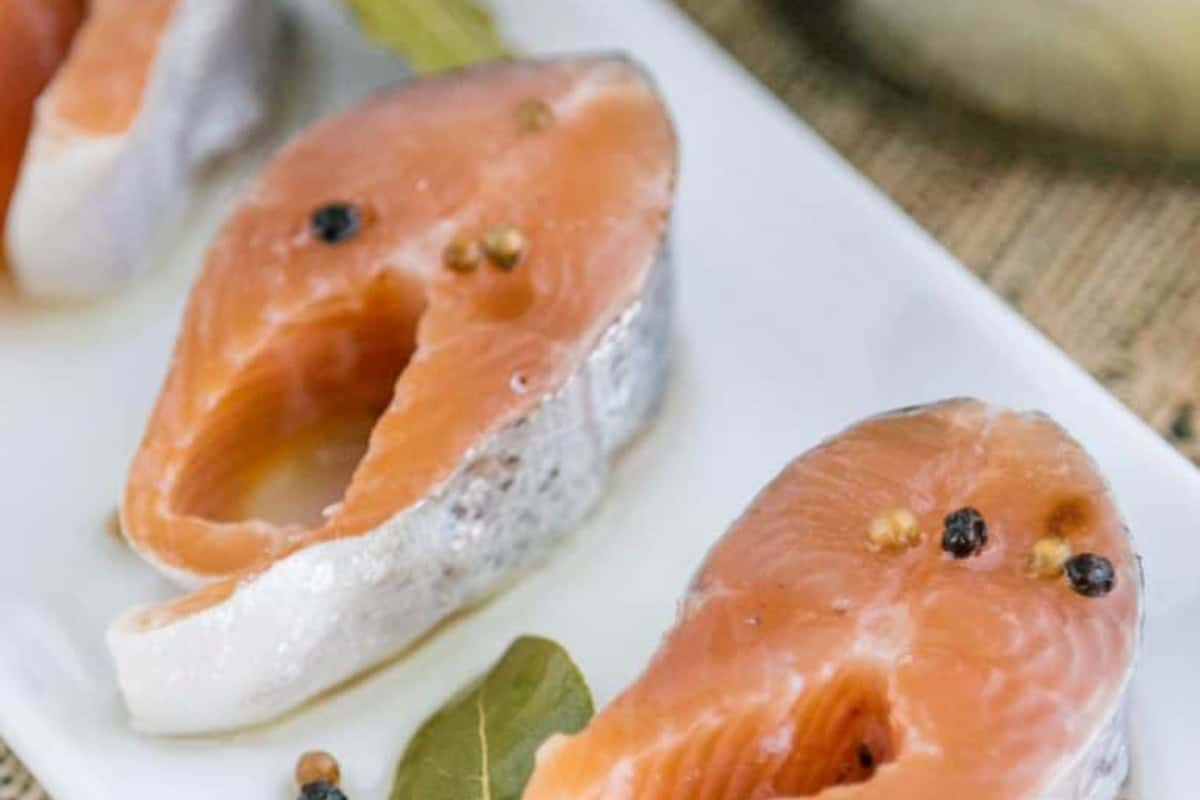 Raw pickled salmon steaks with peppercorns and bay leaves arranged on a white plate.