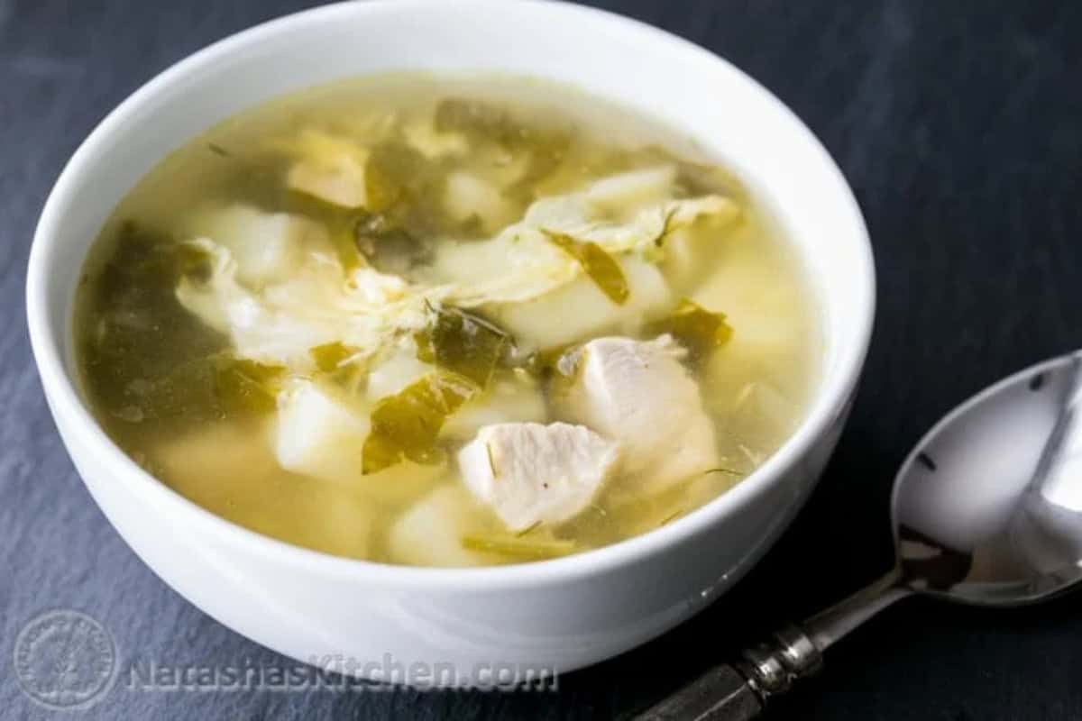 A white bowl of clear chicken sorrel soup with leafy sorrel greens and chunks of chicken placed next to a metal spoon on a dark surface.