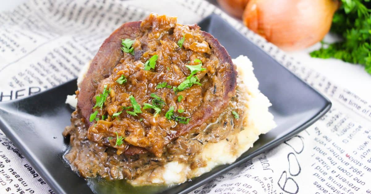 A slice of Smothered Steak and Gravy topped with onion gravy and chopped parsley rests on mashed potatoes, served on a black square plate with onions and herbs in the background.