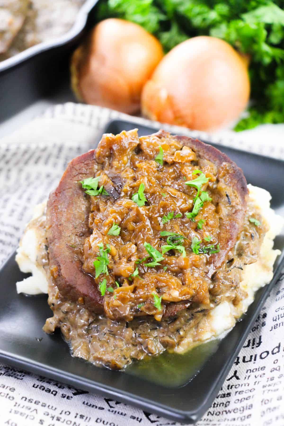 A slice with caramelized onions and herbs sits on mashed potatoes with a creamy sauce, served on a black plate. Onions and leafy greens are in the background.