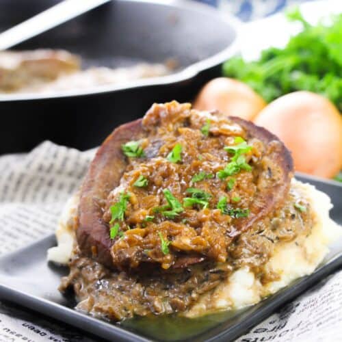 A slice of meatloaf with brown onion gravy sits on mashed potatoes, reminiscent of classic Smothered Steak and Gravy, garnished with chopped parsley on a black plate, with a skillet and onions in the background.