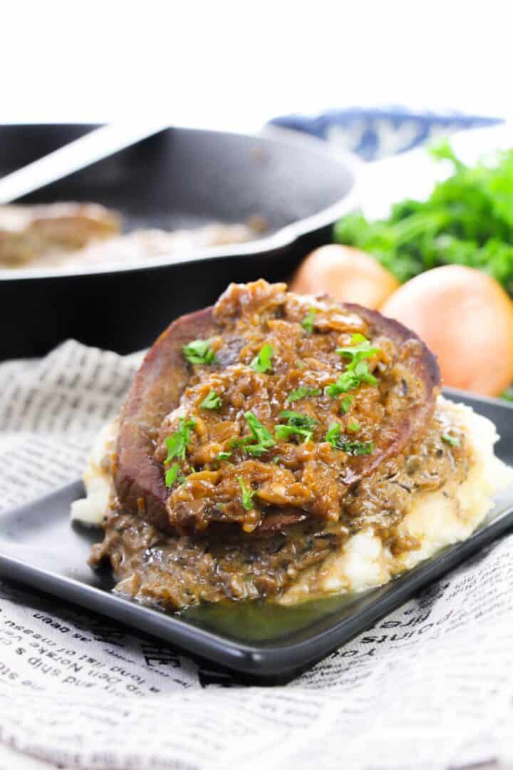 A slice of meatloaf with brown onion gravy sits on mashed potatoes, reminiscent of classic Smothered Steak and Gravy, garnished with chopped parsley on a black plate, with a skillet and onions in the background.