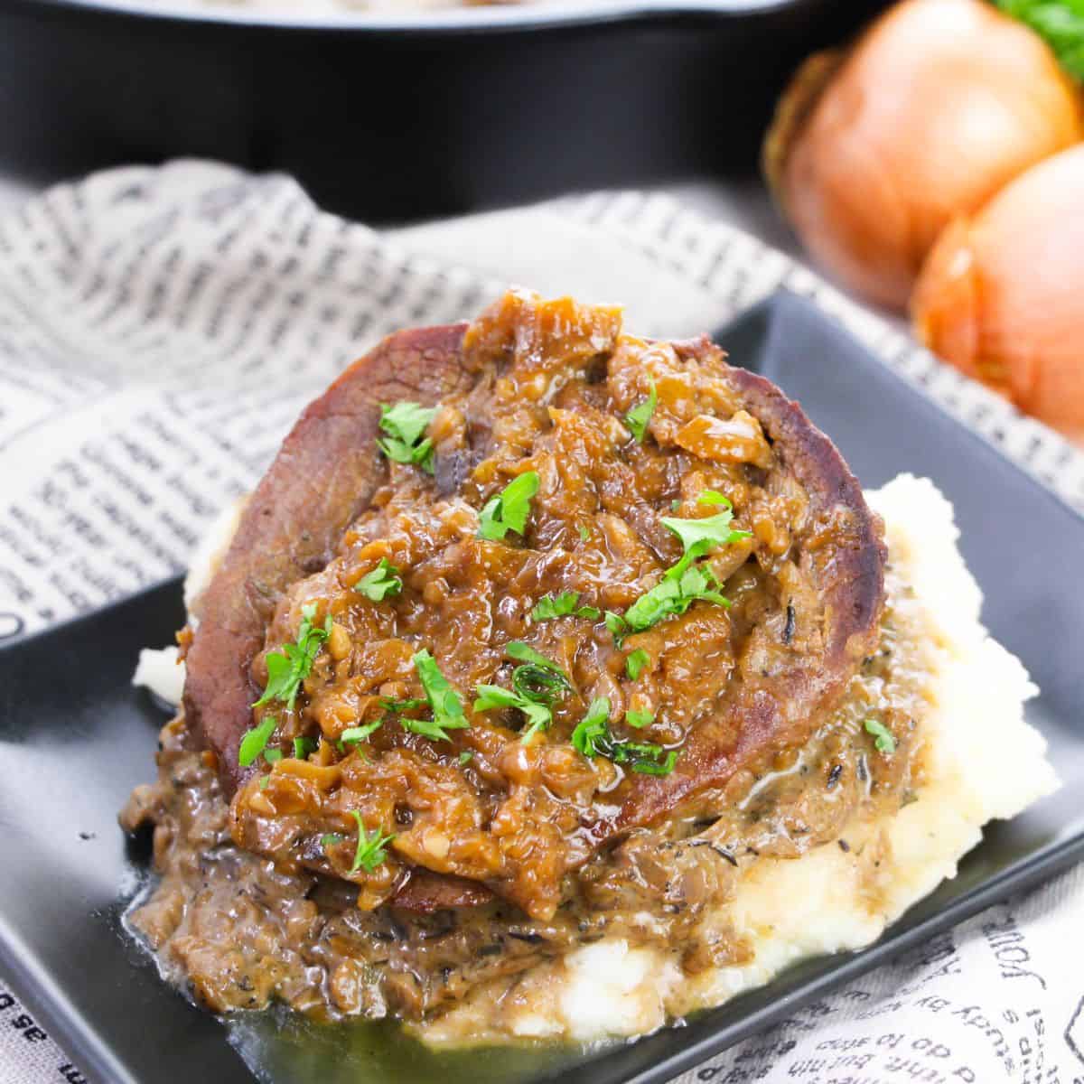 A serving of Smothered Steak and Gravy sits on mashed potatoes, topped with onion gravy and garnished with chopped parsley on a black plate.