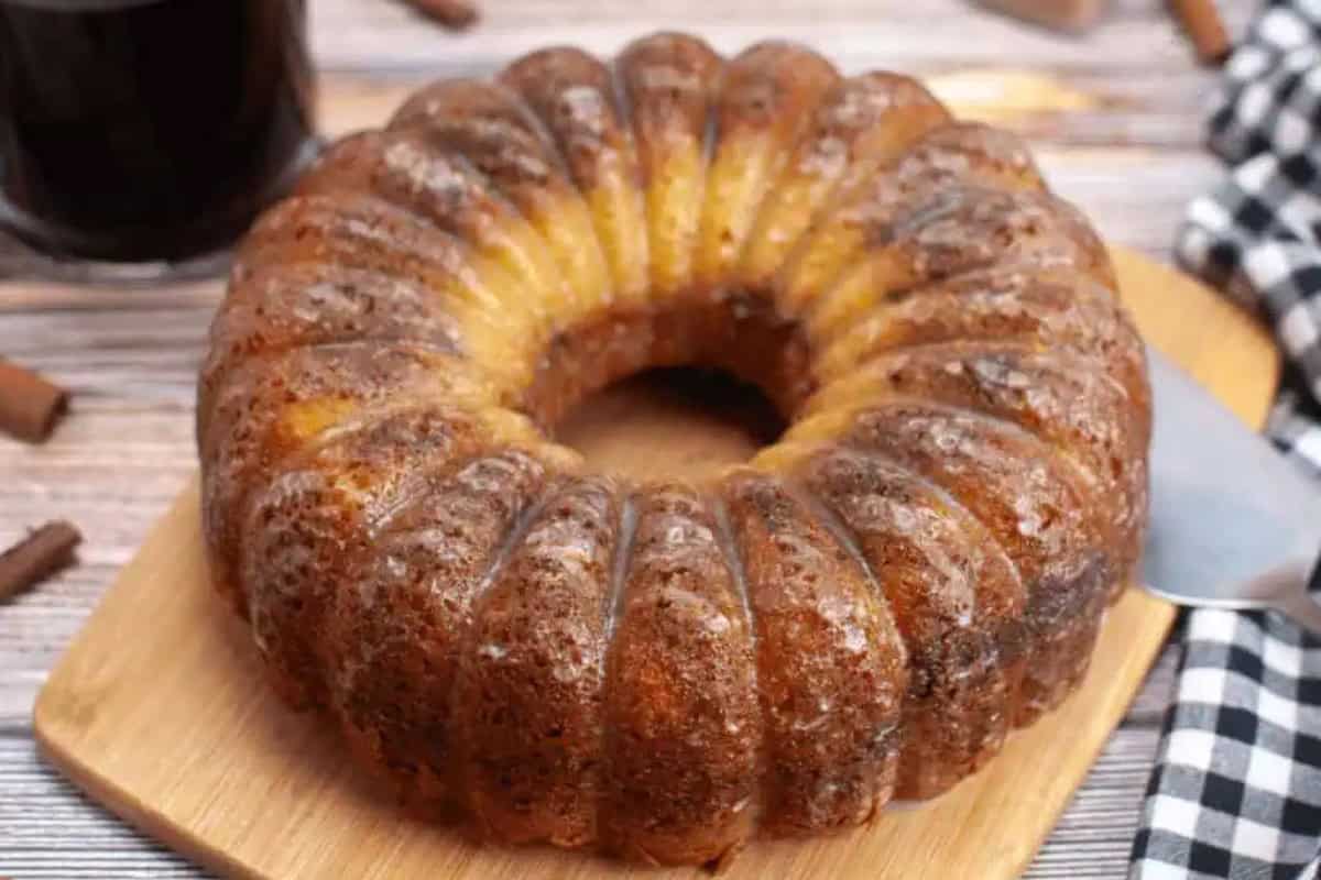A round sour cream bundt coffee cake with a glossy, golden-brown crust sits on a wooden board, next to a knife and a checkered cloth.