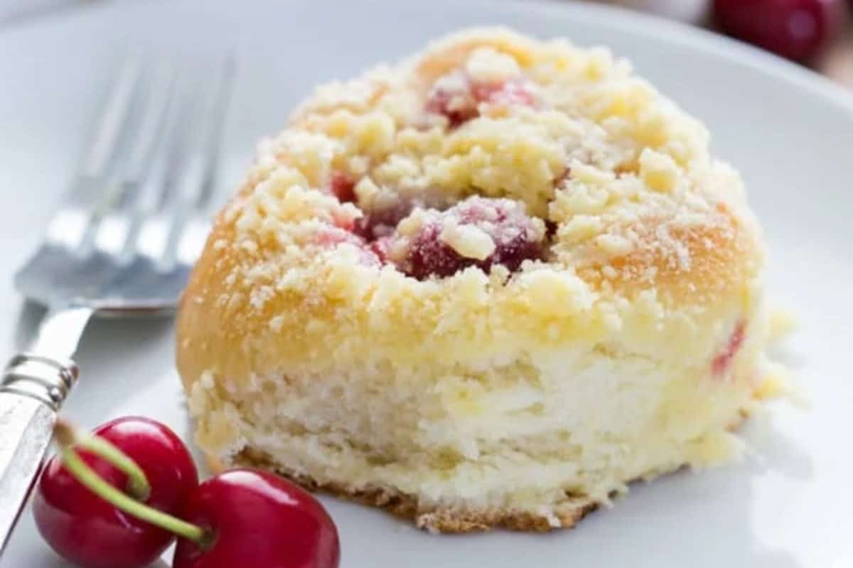 A cherry vatrushka bun, a classic Russian Christmas food, is topped with crumbly streusel and whole cherries, served on a white plate with a fork and fresh cherries.