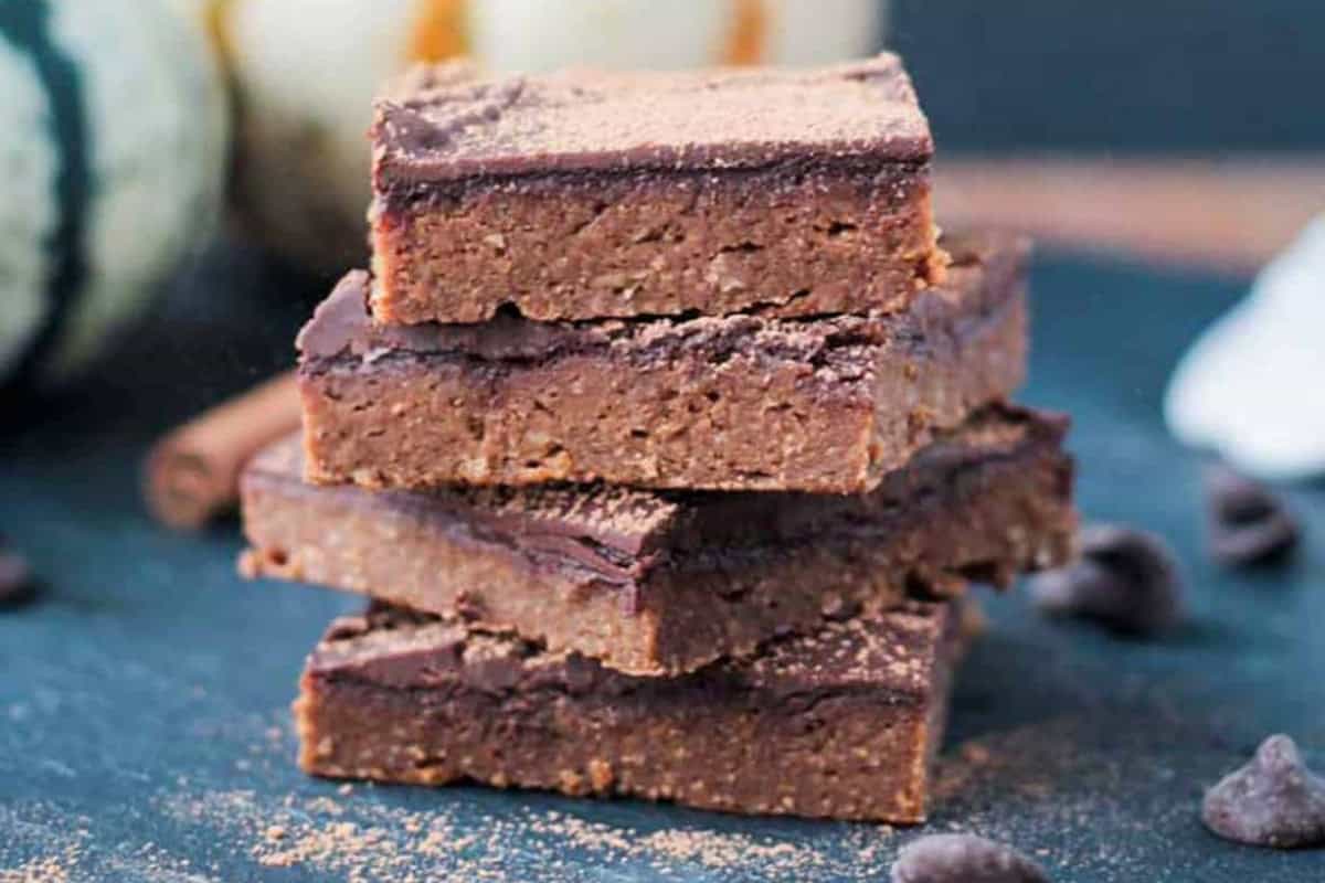 A stack of four vegan pumpkin fudge bars sits on a dark blue surface, surrounded by chocolate chips and a cinnamon stick in the background.
