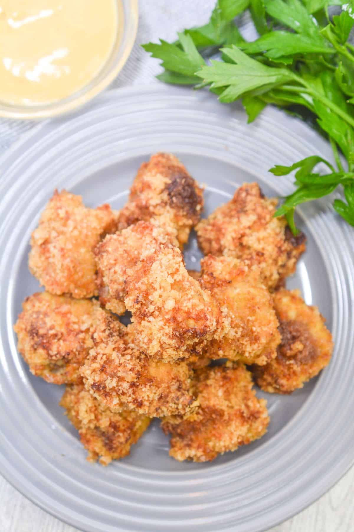 A gray plate with several pieces of breaded, baked  nuggets next to a bunch of fresh parsley and a small bowl of dipping sauce—perfect alongside crispy Air Fryer Steak Bites for a delicious and easy meal.