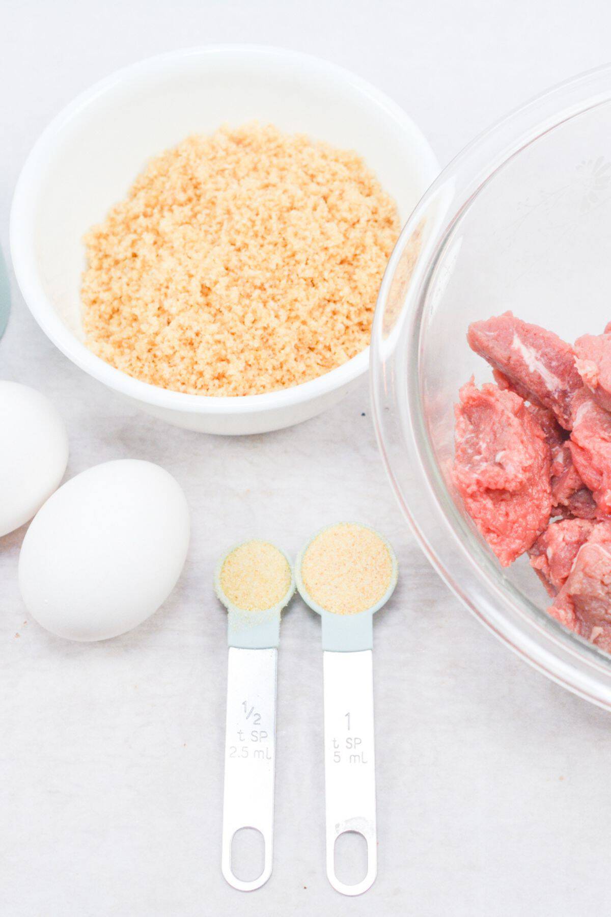 Two eggs, a bowl of breadcrumbs, a bowl of raw meat and measuring spoons with powders are arranged on a white surface.