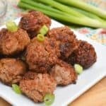 A white plate of Asian Pork Meatballs garnished with chopped green onions, with a bowl of dipping sauce and whole green onions in the background.