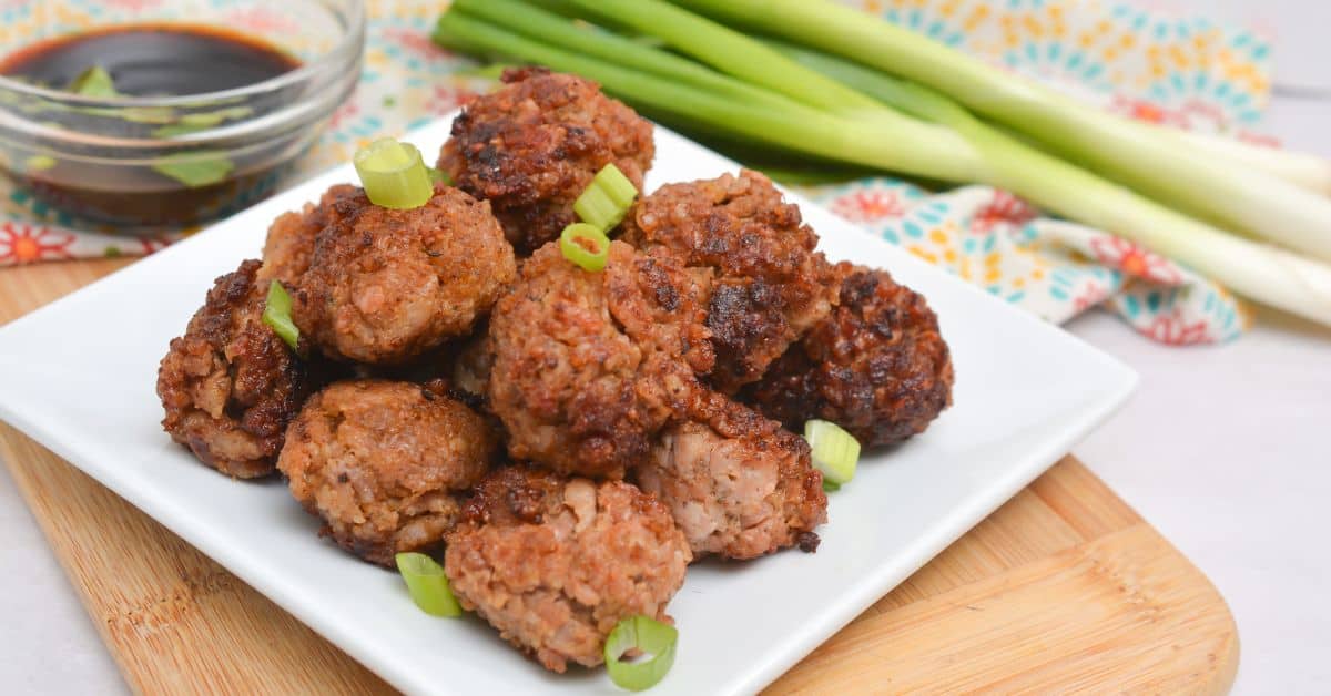 A white plate of Asian Pork Meatballs garnished with chopped green onions, with a bowl of dipping sauce and whole green onions in the background.