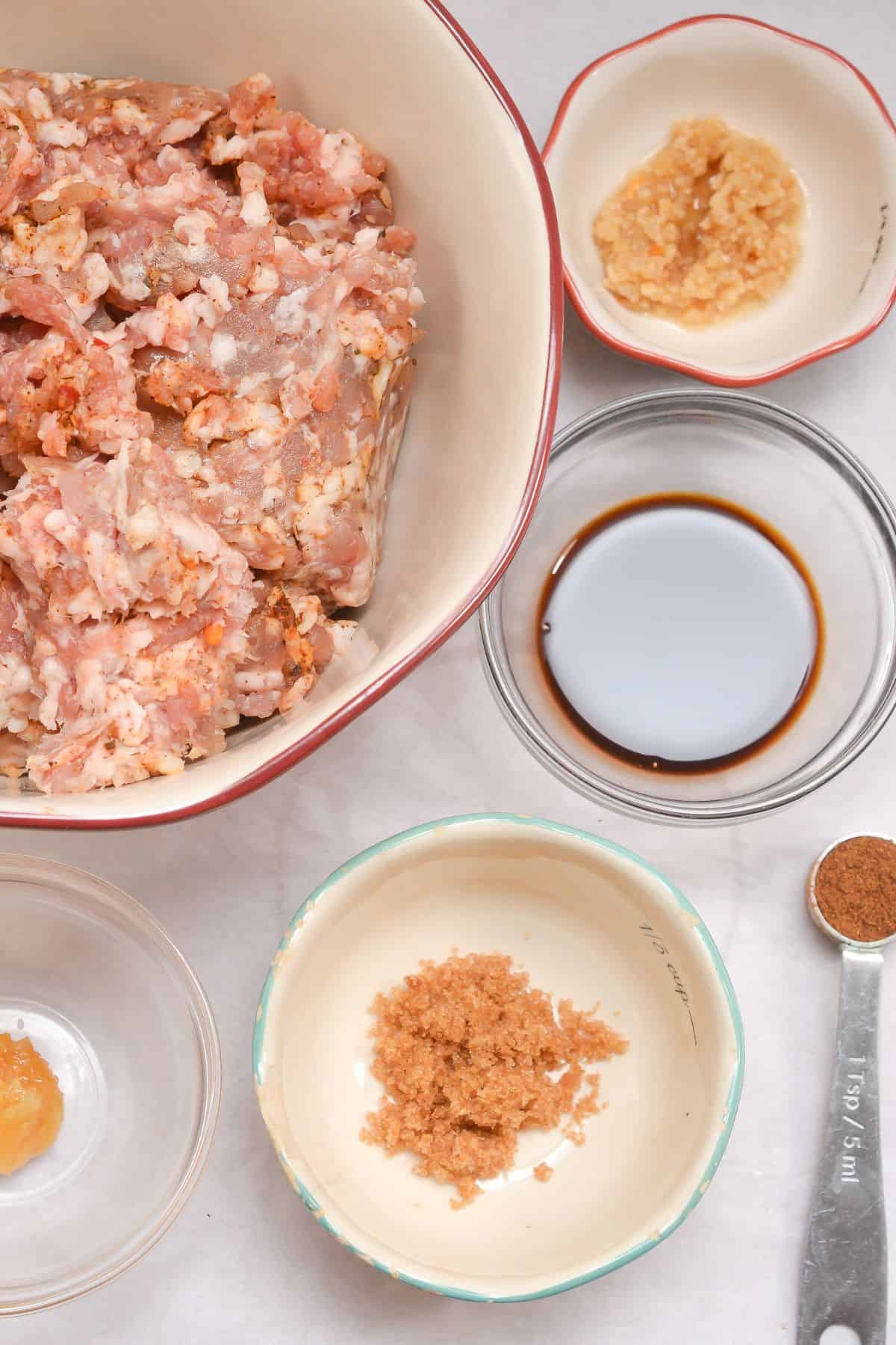 Ground meat in a bowl surrounded by small bowls containing minced garlic, ginger, soy sauce, brown sugar, and a pinch of spice on a white surface—these are the essential ingredients for making flavorful Asian Pork Meatballs.