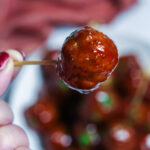 A person holding a barbecue and grape jelly meatball on a toothpick.