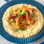 A blue plate with creamy grits topped with Cajun Shrimp and Grits, sautéed shrimp, and chopped green onions, served on a white textured surface.