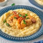 A blue plate with creamy grits topped with Cajun Shrimp and Grits, garnished with chopped green onions; side dishes in small bowls in the background.