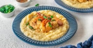 A blue plate with creamy grits topped with Cajun Shrimp and Grits, garnished with chopped green onions; side dishes in small bowls in the background.