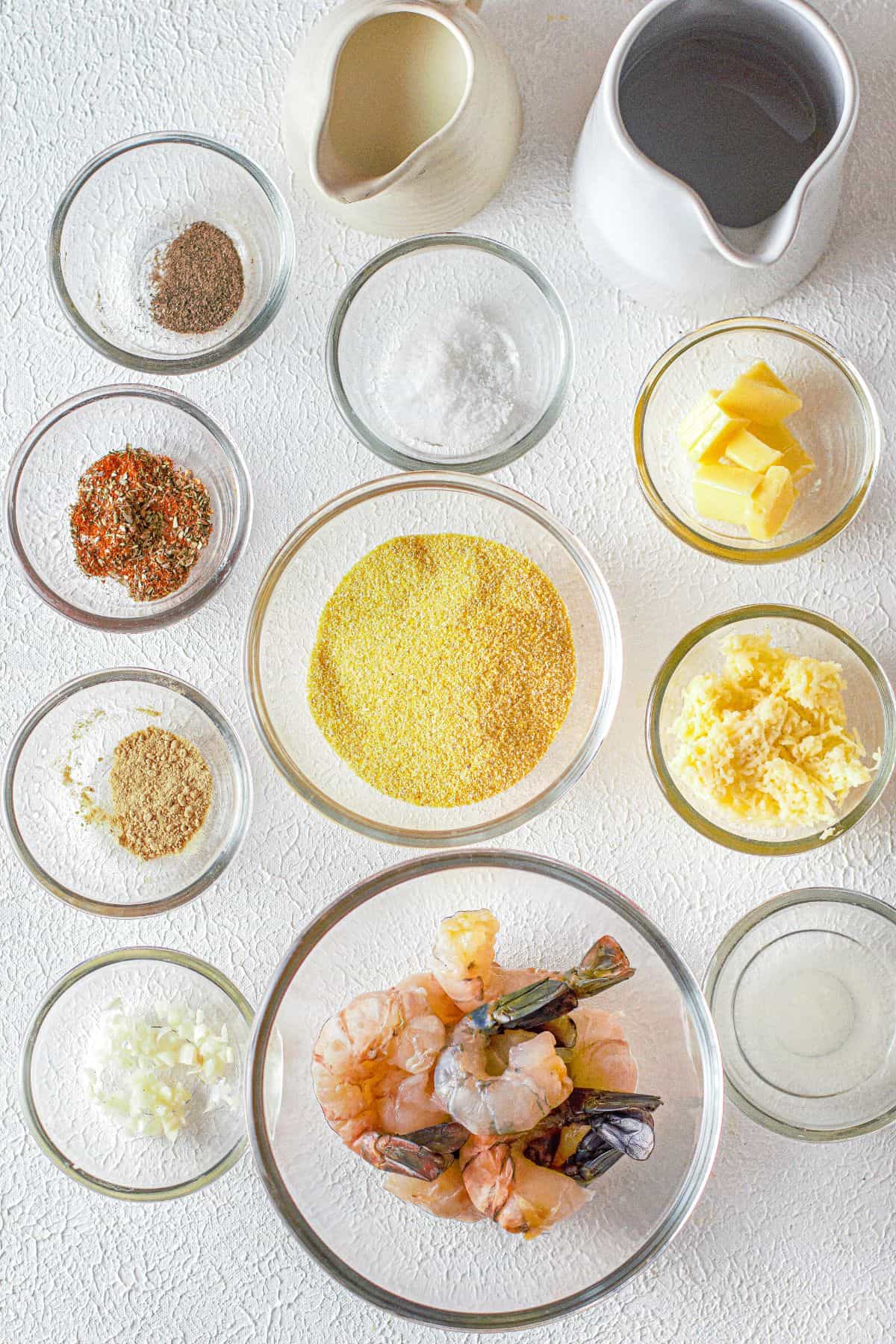 Various ingredients in glass bowls on a white surface, including raw shrimp, cornmeal, shredded cheese, butter, spices, chopped onions, flour, water, and milk—perfect for making classic Cajun Shrimp and Grits.