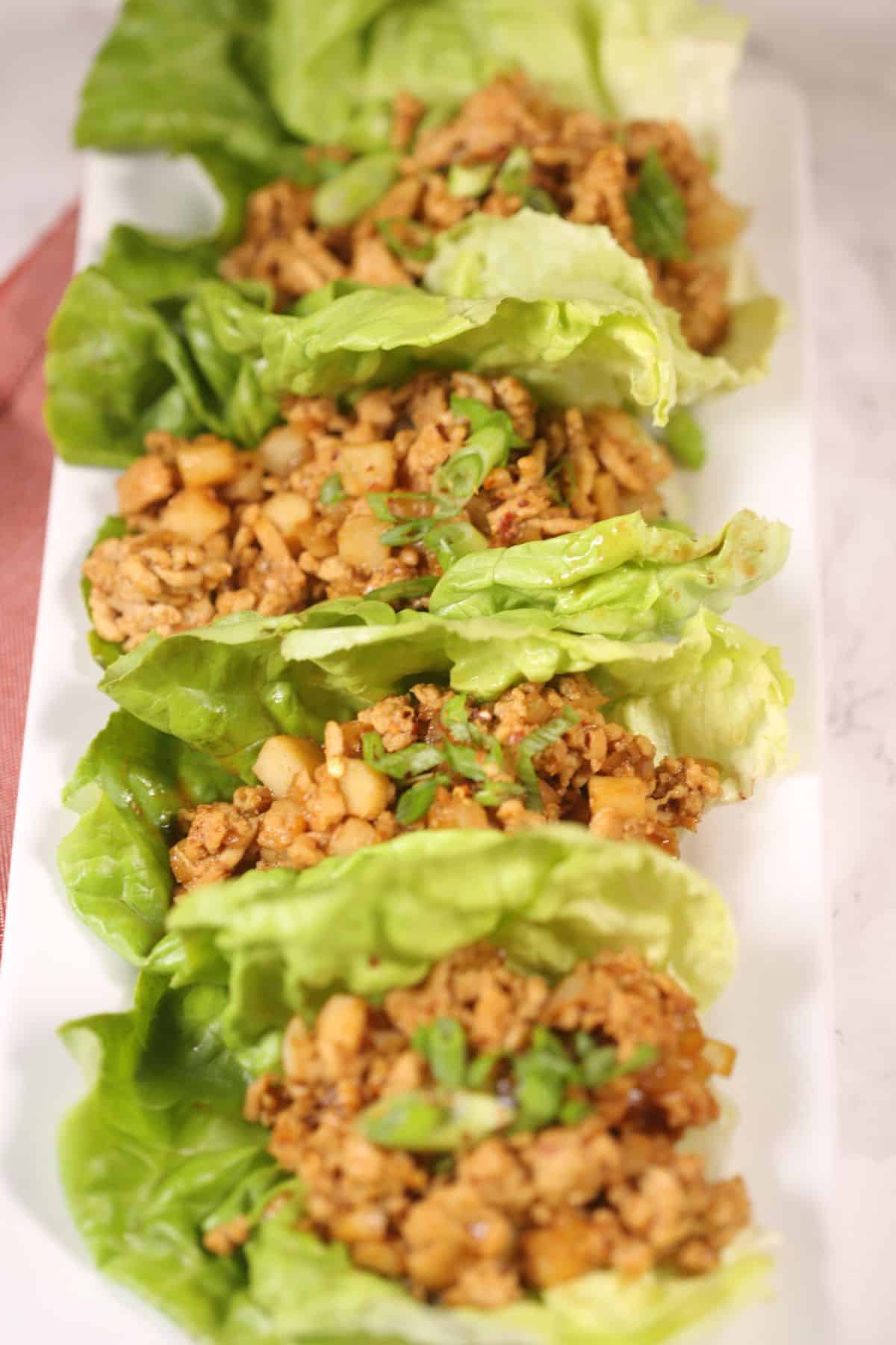Four Asian Lettuce Wraps filled with seasoned ground meat, diced vegetables, and chopped green onions, arranged neatly on a white rectangular plate.