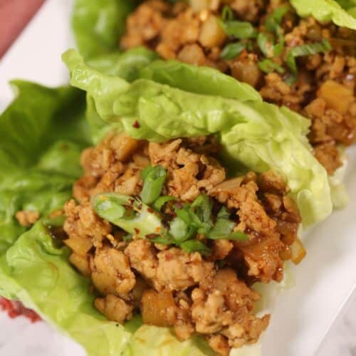 A close-up of Asian Lettuce Wraps filled with cooked ground meat, diced vegetables, and garnished with chopped green onions on a white plate.