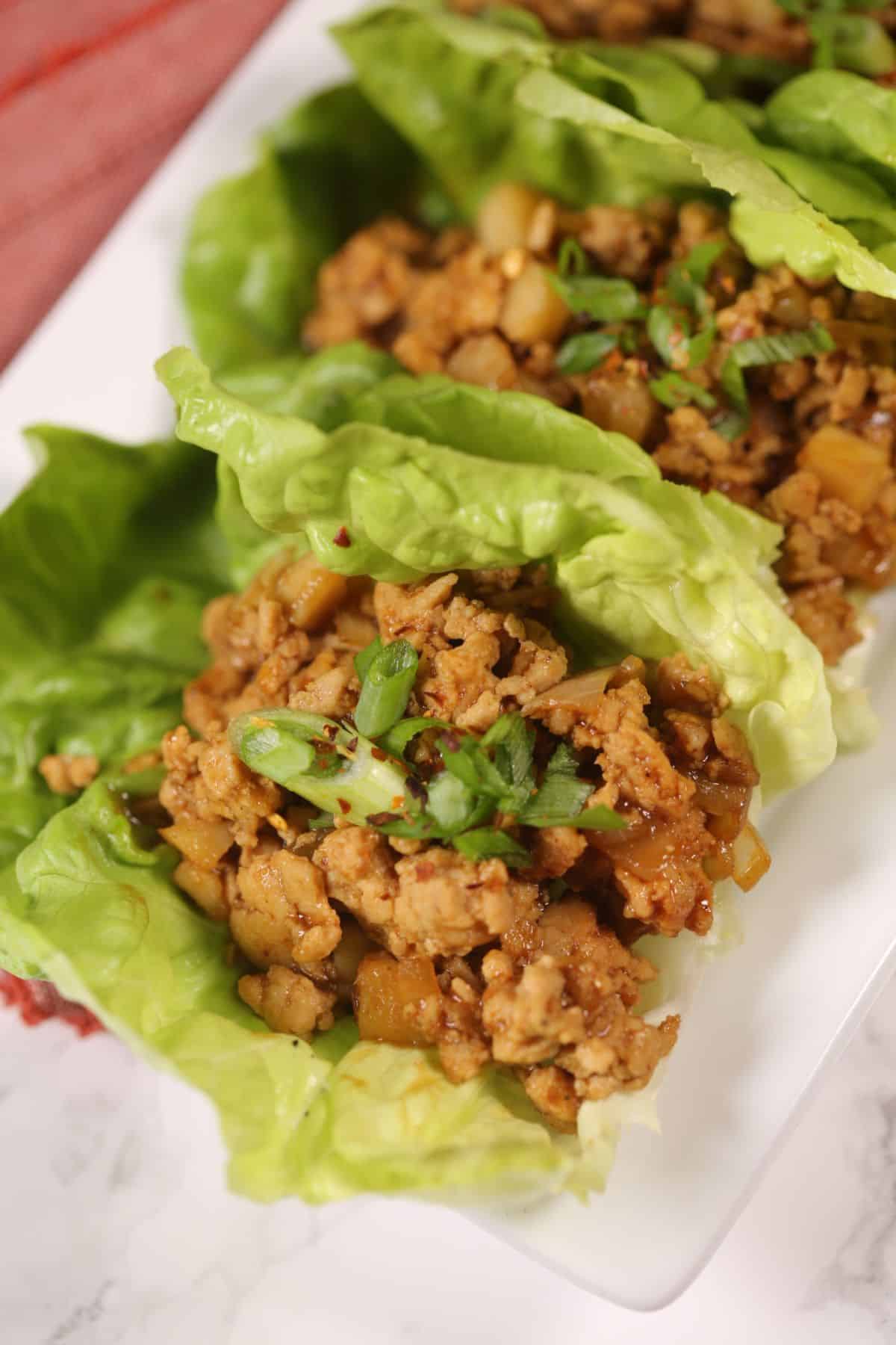 A close-up of Asian Lettuce Wraps filled with cooked ground meat, diced vegetables, and garnished with chopped green onions on a white plate.