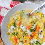 A bowl of Chicken Corn Noodle Soup with carrots, parsley, and egg noodles, served steaming hot with a spoon and a slice of bread on the side.