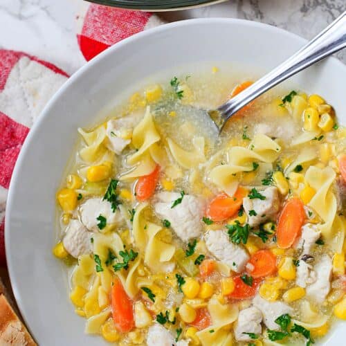 A bowl of Chicken Corn Noodle Soup with carrots, parsley, and egg noodles, served steaming hot with a spoon and a slice of bread on the side.