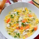 A white bowl filled with Chicken Corn Noodle Soup, featuring egg noodles, carrots, corn, and herbs, served with a spoon and bread on the side.