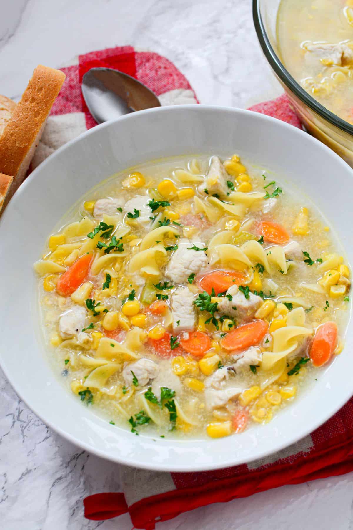 A white bowl featuring egg noodles, carrots, and herbs, served with a spoon and bread on the side.