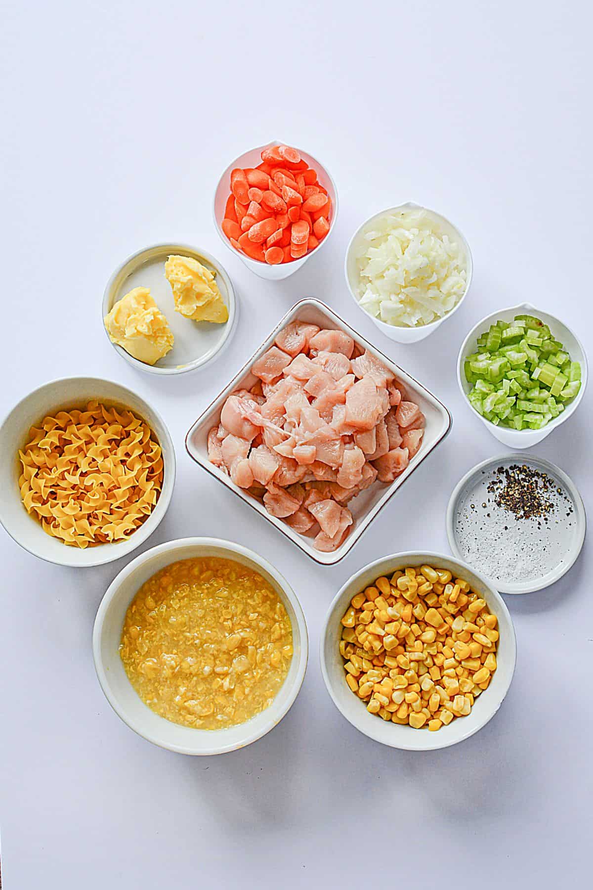 Nine bowls display the key ingredients : raw diced meat, chopped carrots, onion, celery, corn, egg noodles, butter, a beaten egg mixture, and salt and pepper arranged on a white background.