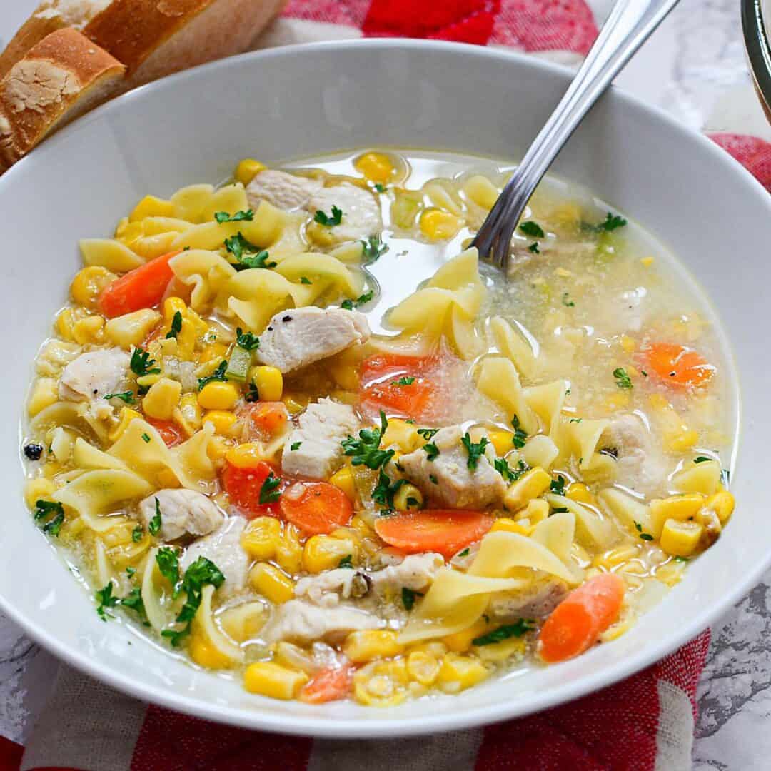 A bowl of Chicken Corn Noodle Soup with carrots, parsley, and wide noodles, served with a spoon and a slice of bread on the side.