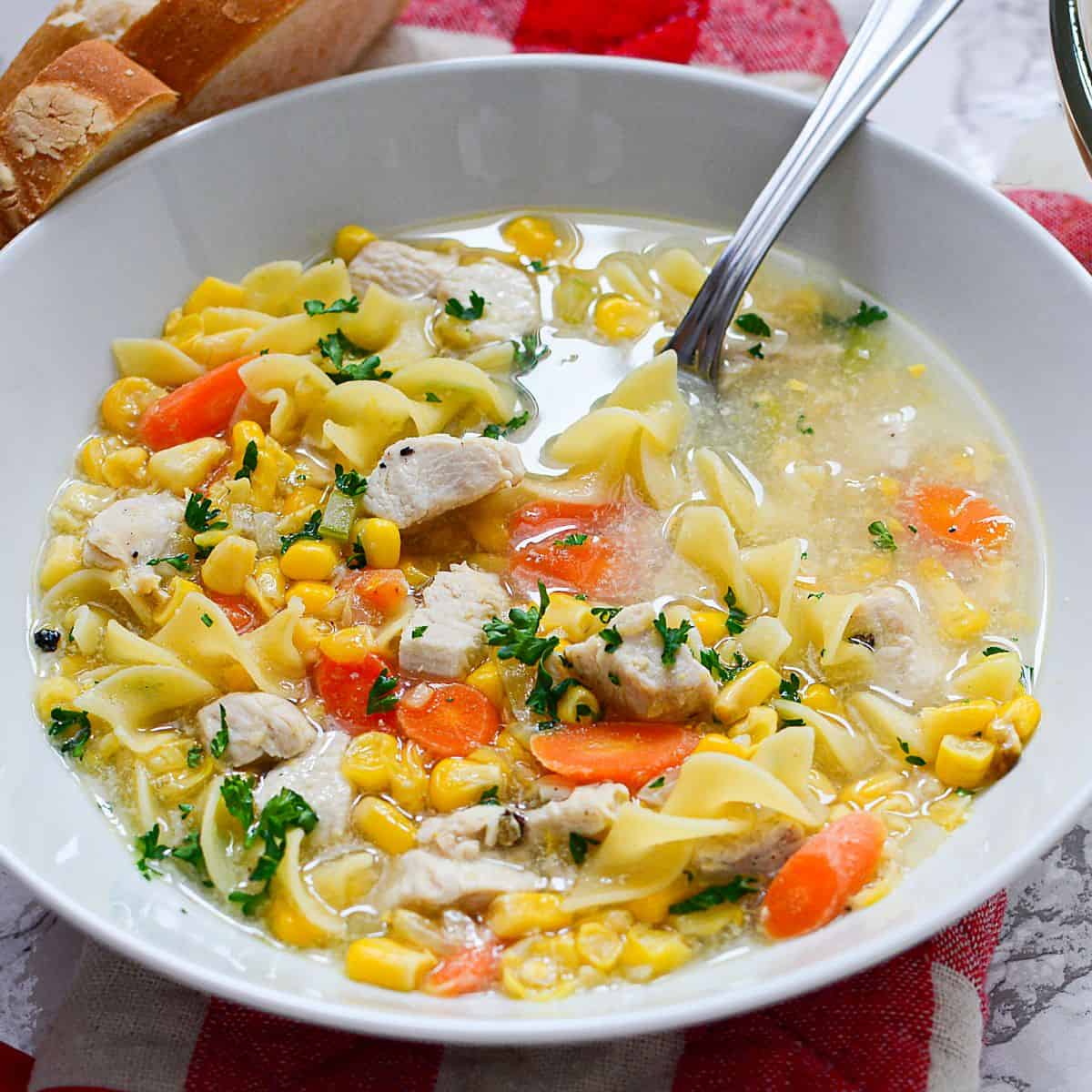 A bowl of Chicken Corn Noodle Soup with carrots, parsley, and wide noodles, served with a spoon and a slice of bread on the side.