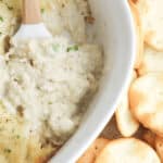 A white dish filled with a rich Cream Cheese and Crab Dip is next to a small pile of round crackers. A spatula rests in the dip.