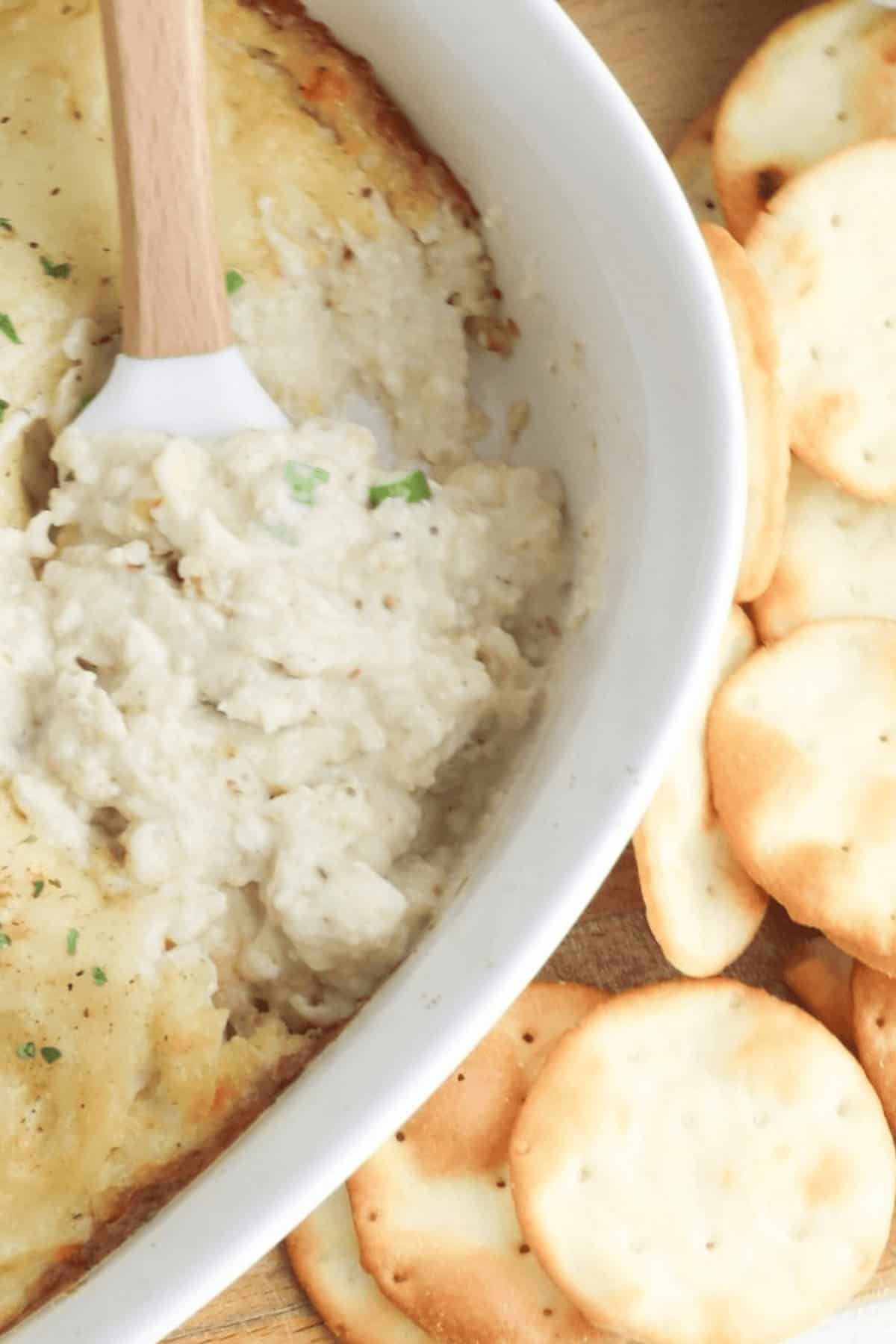 A white dish filled with a rich Cream Cheese and Crab Dip is next to a small pile of round crackers. A spatula rests in the dip.