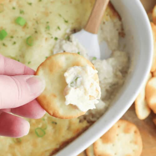 A hand holds a cracker with Cream Cheese and Crab Dip, poised above a white dish brimming with the creamy baked delight. Chopped green onions adorn the top, while a wooden spoon peeks from the side.