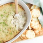 A white dish brimming with baked cream cheese and crab dip, garnished with fresh green onions. Nestled alongside is a wooden spoon and a heap of round crackers on a rustic wooden board.
