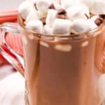 A clear glass mug filled with Slow Cooker Hot Cocoa, topped with mini marshmallows and a few chocolate chips, sits on a white plate.