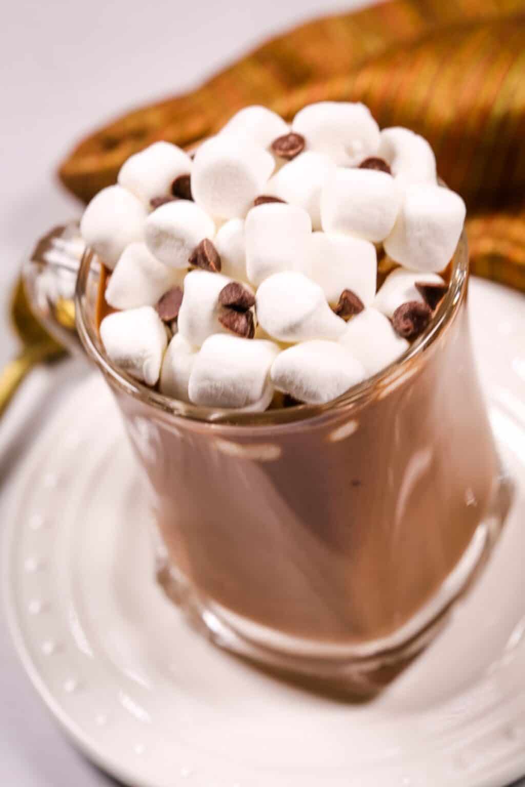 A glass mug of Slow Cooker Hot Cocoa topped with mini marshmallows and chocolate chips, placed on a white saucer with a spoon and a striped cloth nearby.