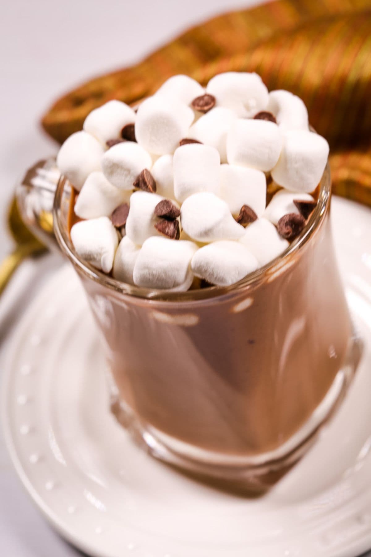 A glass mug of Slow Cooker Hot Cocoa topped with mini marshmallows and chocolate chips, placed on a white saucer with a spoon and a striped cloth nearby.