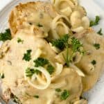 Two pork chops with mushrooms and gravy, topped with creamy onion sauce and fresh parsley, served on a white plate.