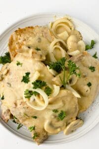 Two pork chops with mushrooms and gravy, topped with creamy onion sauce and fresh parsley, served on a white plate.