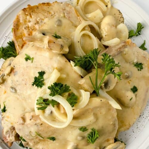 Two pork chops with mushrooms and gravy, topped with creamy onion sauce and fresh parsley, served on a white plate.