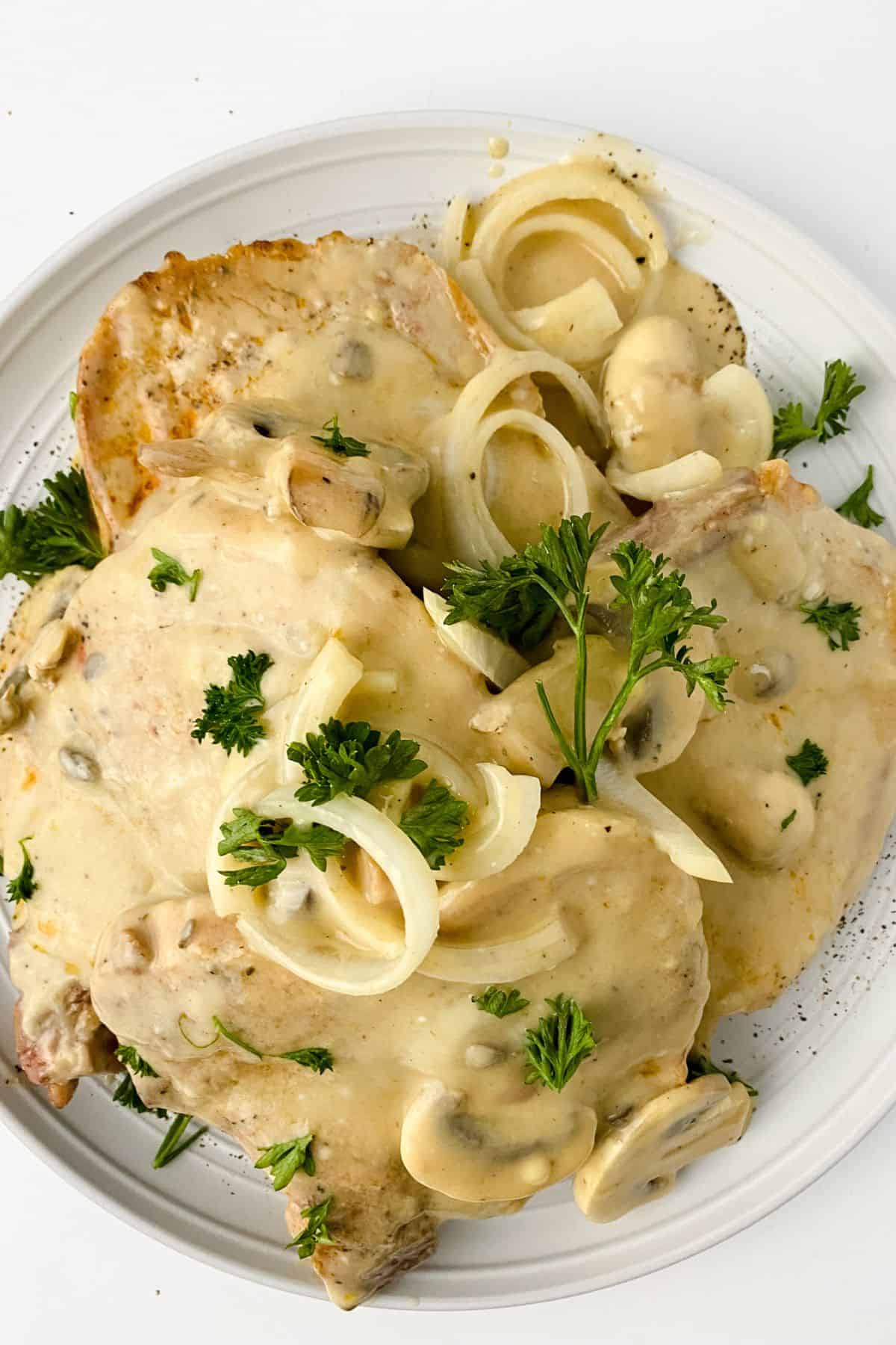 Two pork chops with mushrooms and gravy, topped with creamy onion sauce and fresh parsley, served on a white plate.