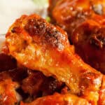 Close-up of several crispy, saucy buffalo and honey BBQ chicken wings with a side of creamy dipping sauce in the background.