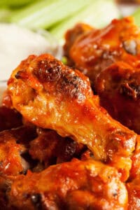 Close-up of several crispy, saucy buffalo and honey BBQ chicken wings with a side of creamy dipping sauce in the background.