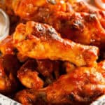 A pile of saucy Honey BBQ wings sits invitingly on a plate, accompanied by a small cup of dipping sauce in the background.