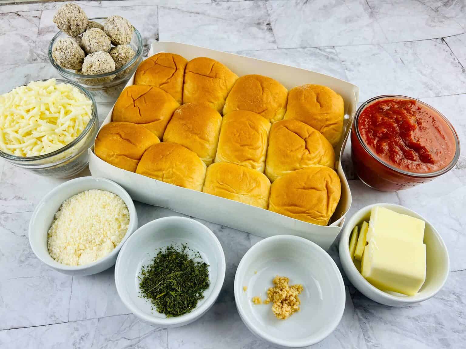 A box of dinner rolls surrounded by bowls brimming with shredded cheese, Parmesan, herbs, garlic, butter, marinara sauce—all you need for crafting.