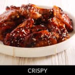 A white plate of crispy Korean BBQ wings garnished with sesame seeds, resting invitingly on a light-colored wooden surface.
