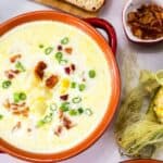 A bowl of sweet corn soup garnished with bacon and green onions, with fresh corn, bread, and a bowl of salt nearby.