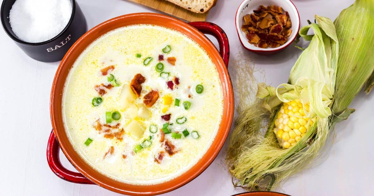 A bowl of sweet corn soup garnished with bacon and green onions, with fresh corn, bread, and a bowl of salt nearby.